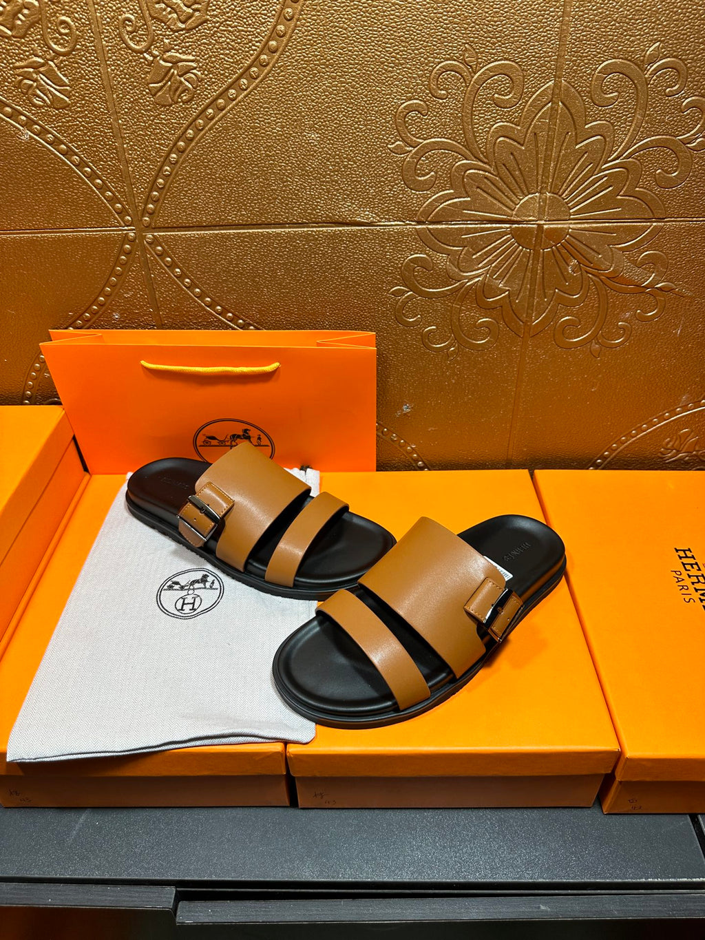 Tan and black Hermes slide sandals on box with white logo cloth