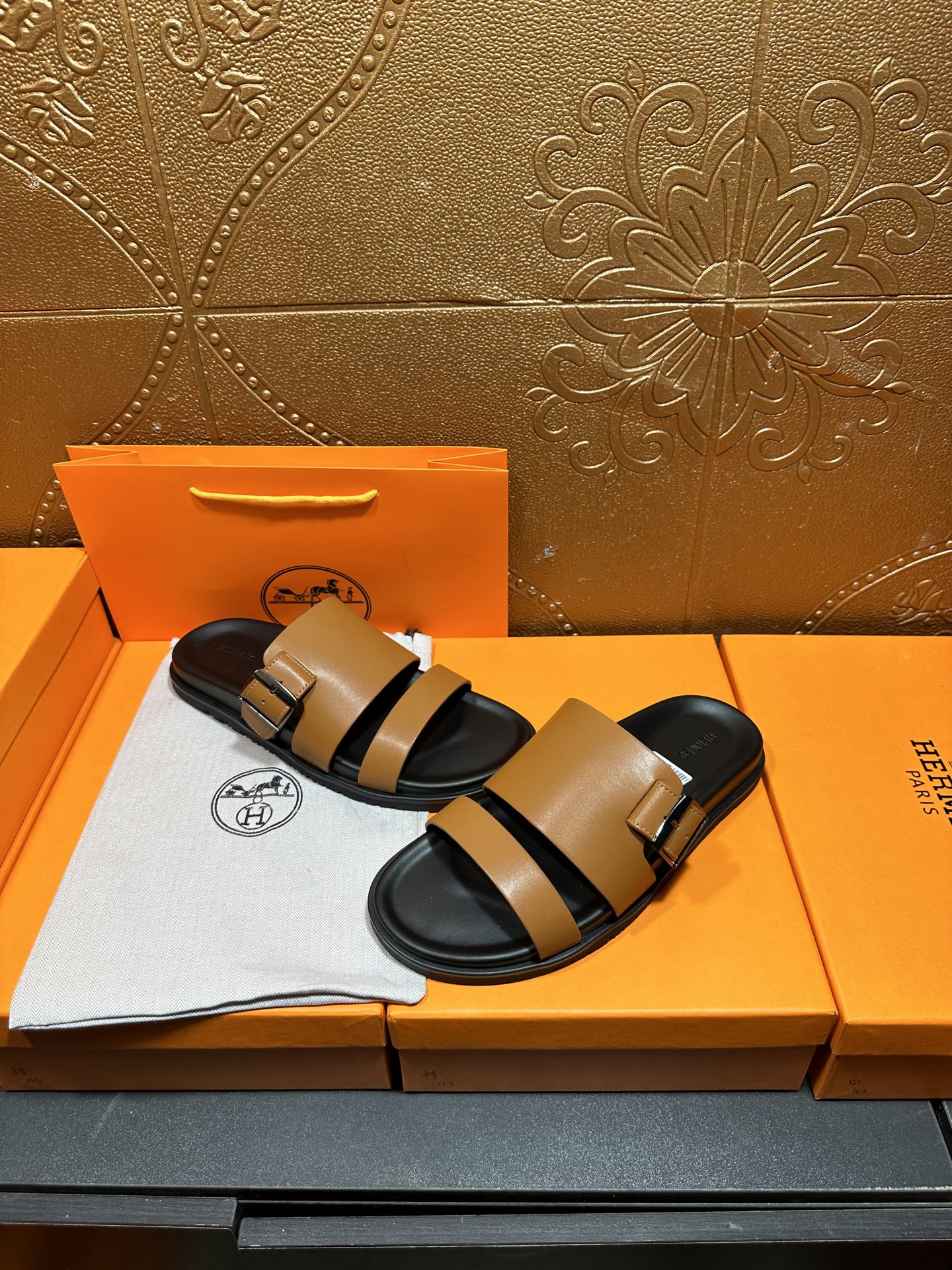 Tan and black Hermes slide sandals on box with white logo cloth