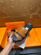 Tan and black Hermes slide sandals on white cloth, hand adjusting strap