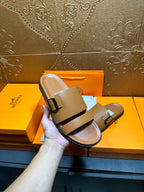 Tan leather slide sandals held in hand, two buckle straps over cushioned footbed