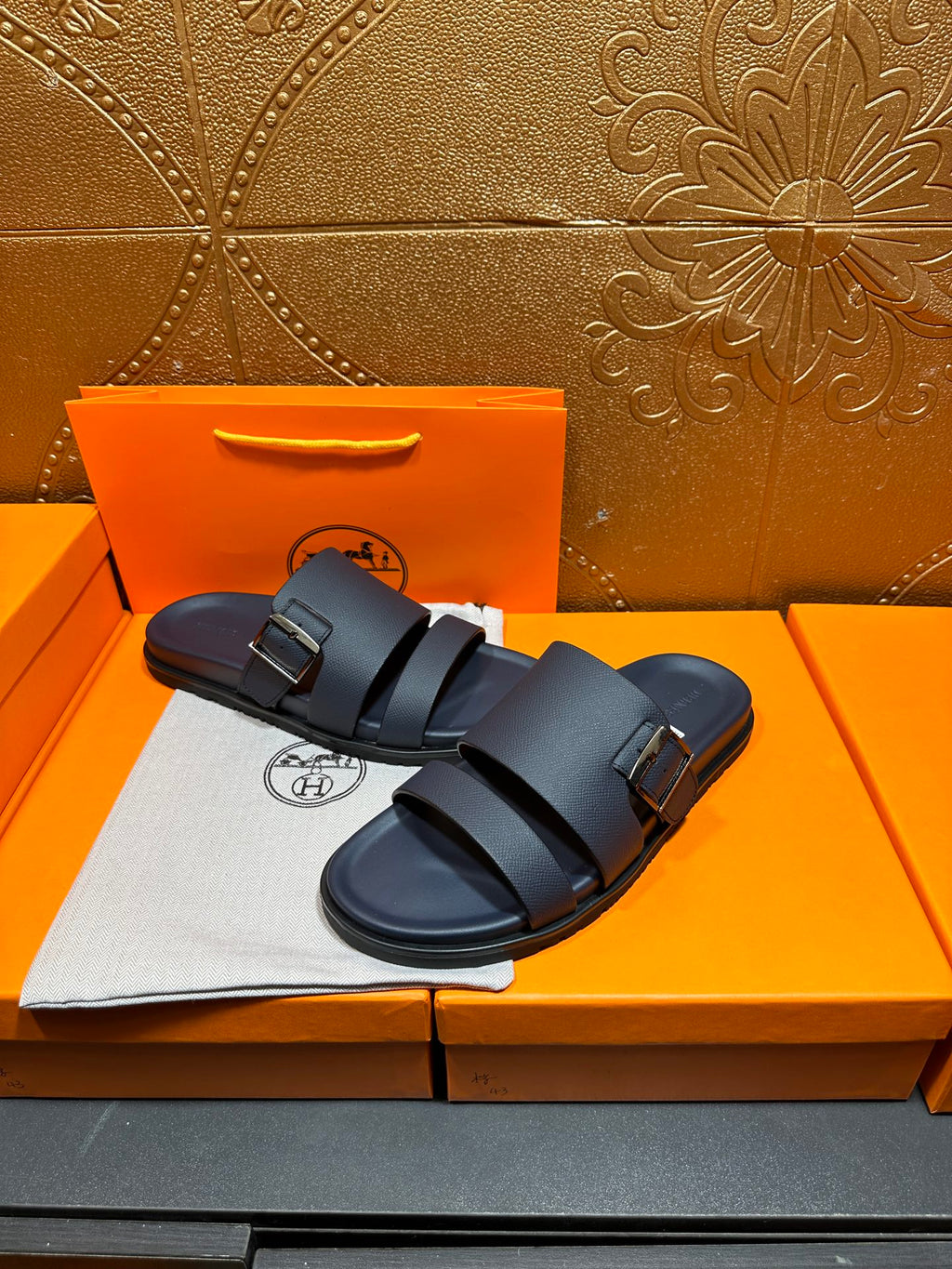 Blue leather slide sandals with double buckled straps, resting on box