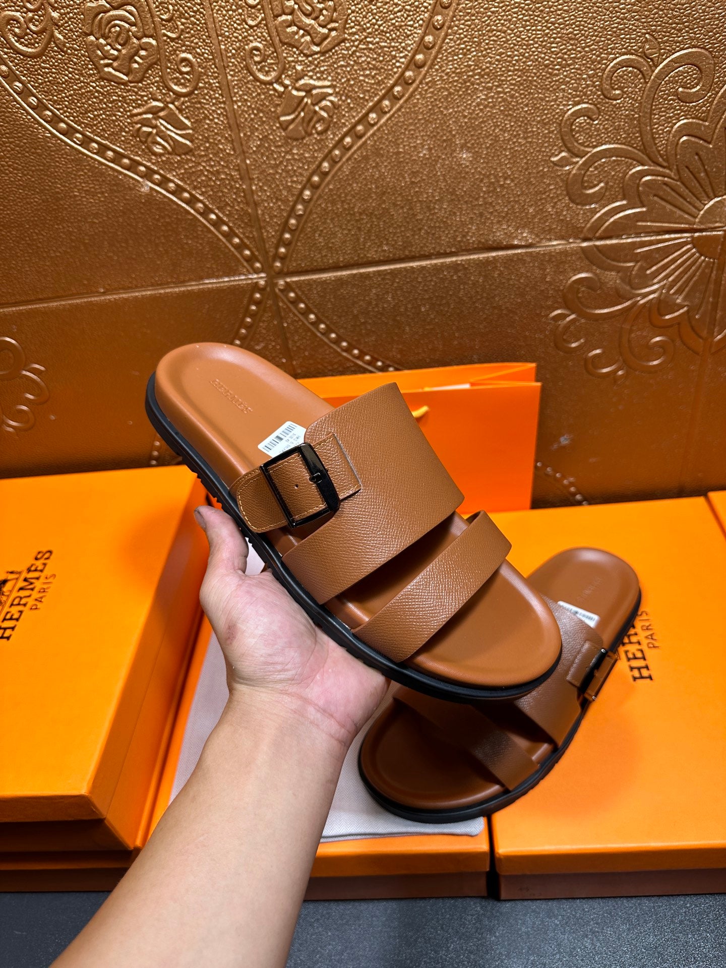 Hermes tan-brown leather sandal held in hand, wide band straps and buckle