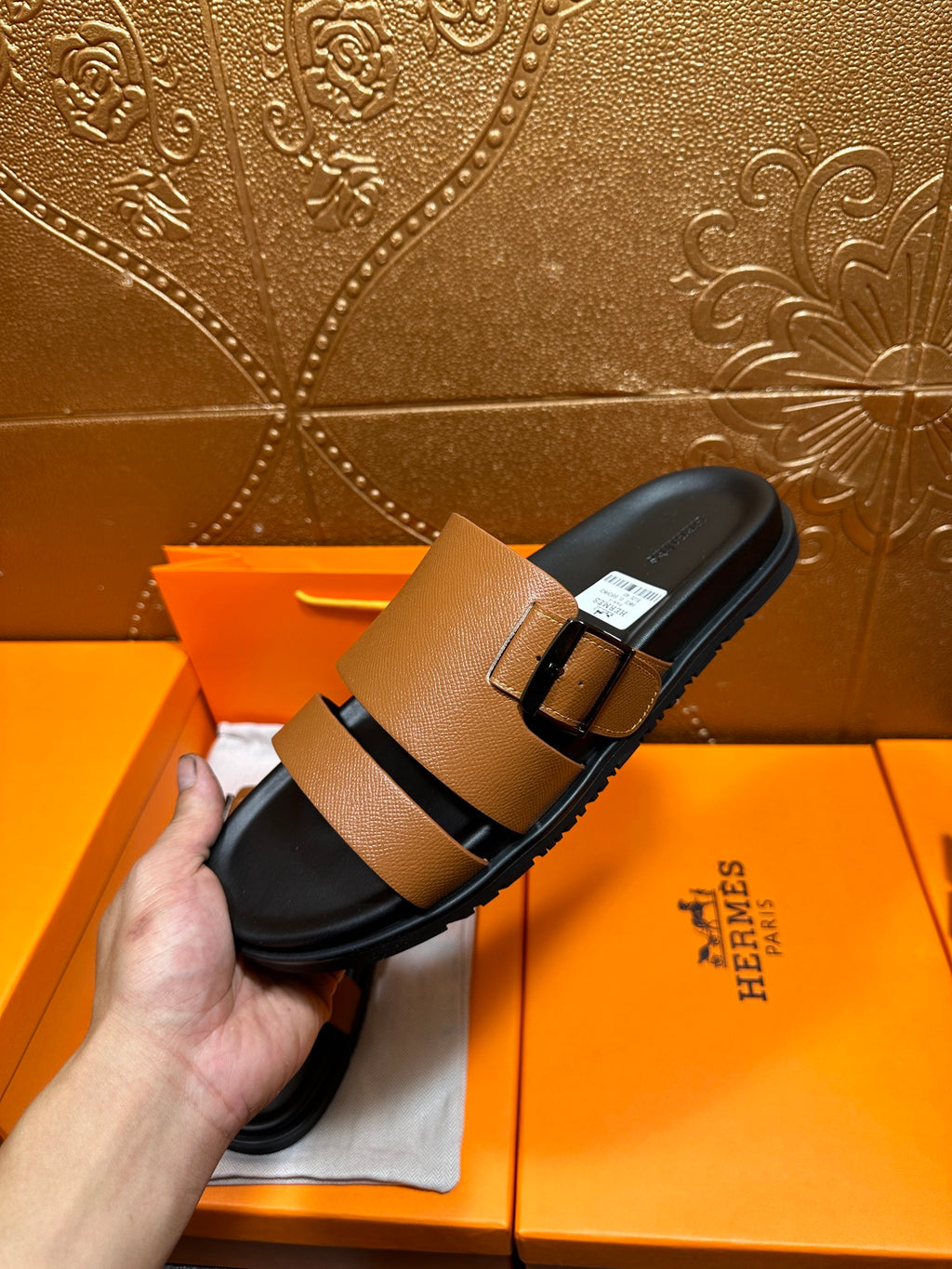 Brown slide sandal with black strap and metal buckle held up in front of packaging.