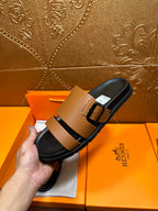 Brown slide sandal with black strap and metal buckle held up in front of packaging.
