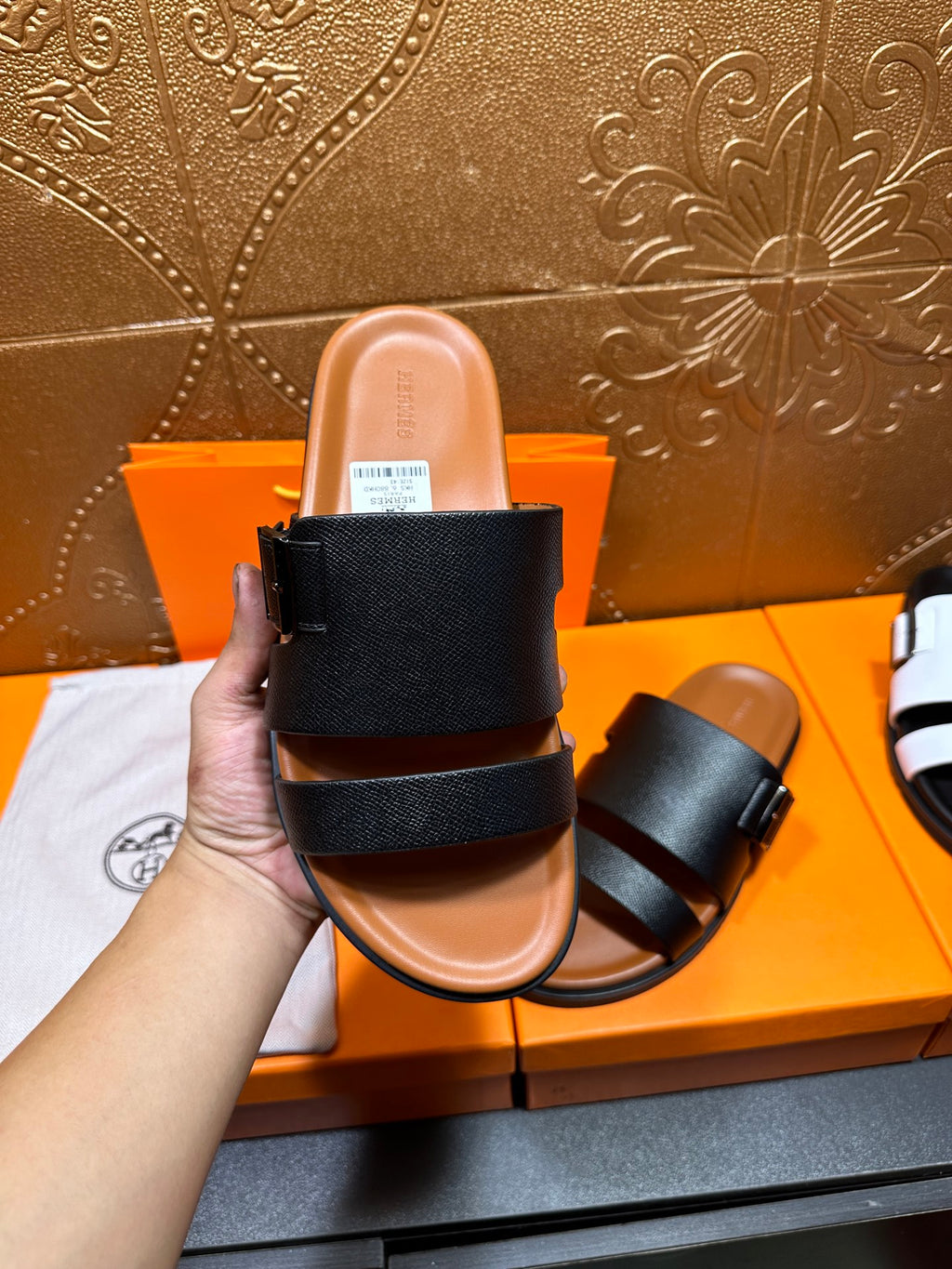 Black slide sandal with two wide straps, hand holds toe opening, tan footbed