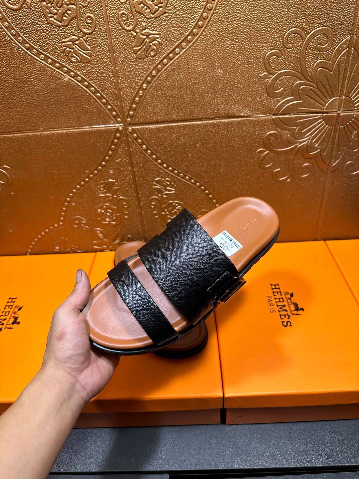 Black slide sandal with two wide straps, hand holds one shoe over box