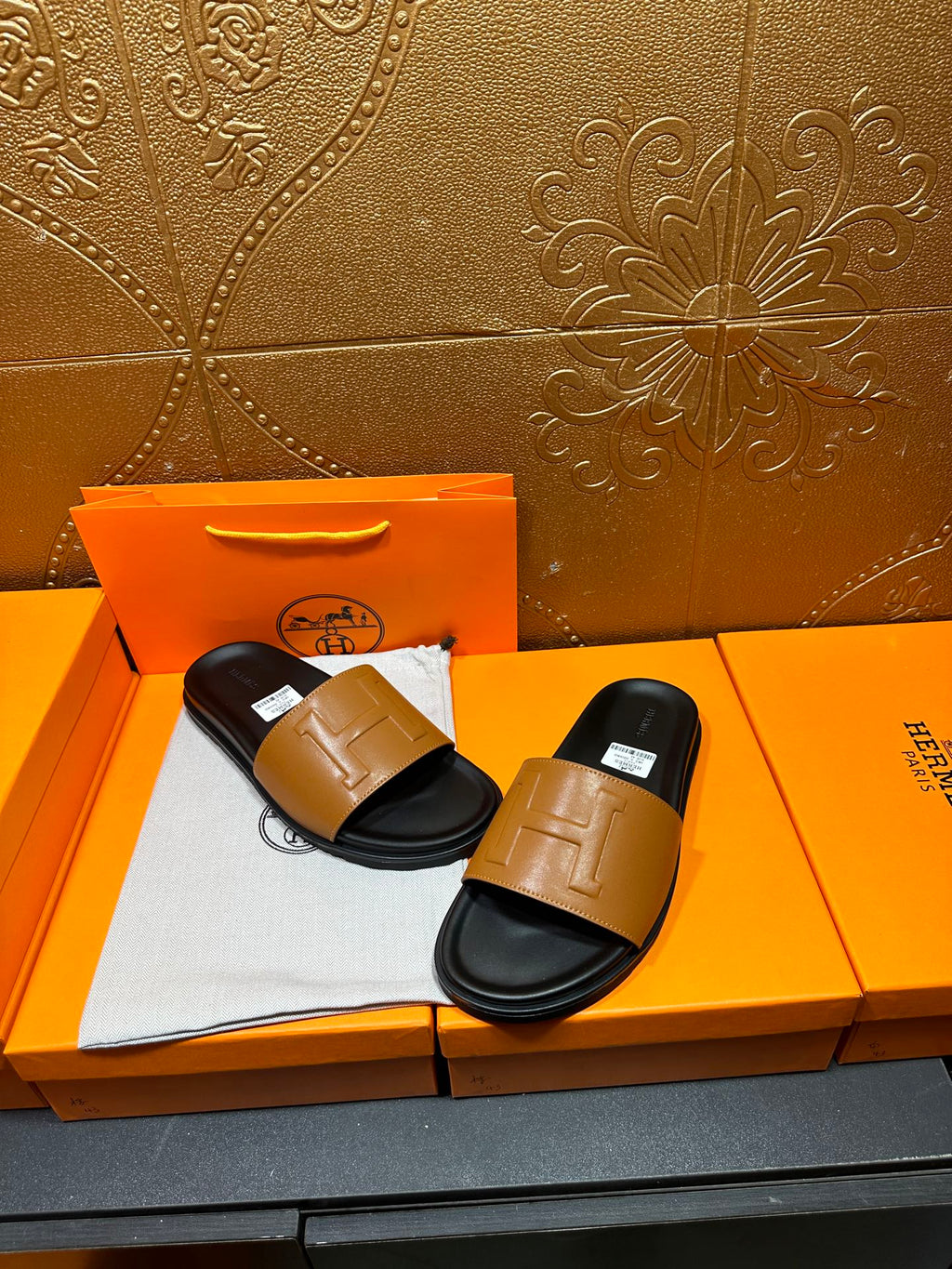 Hermes slide sandals with tan straps on black footbeds, displayed on orange box