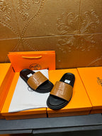 Hermes slide sandals with tan straps on black footbeds, displayed on orange box