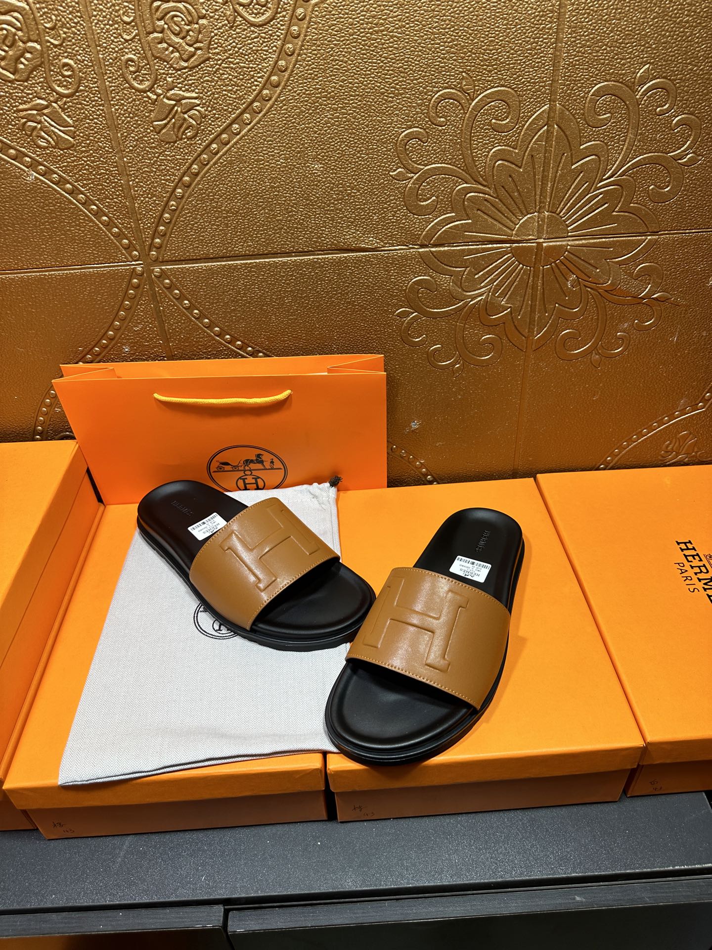 Hermes slide sandals with tan straps on black footbeds, displayed on orange box