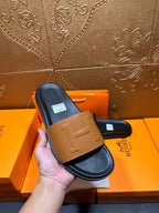 Hermes slide sandals held in hand: tan front strap on dark footbed