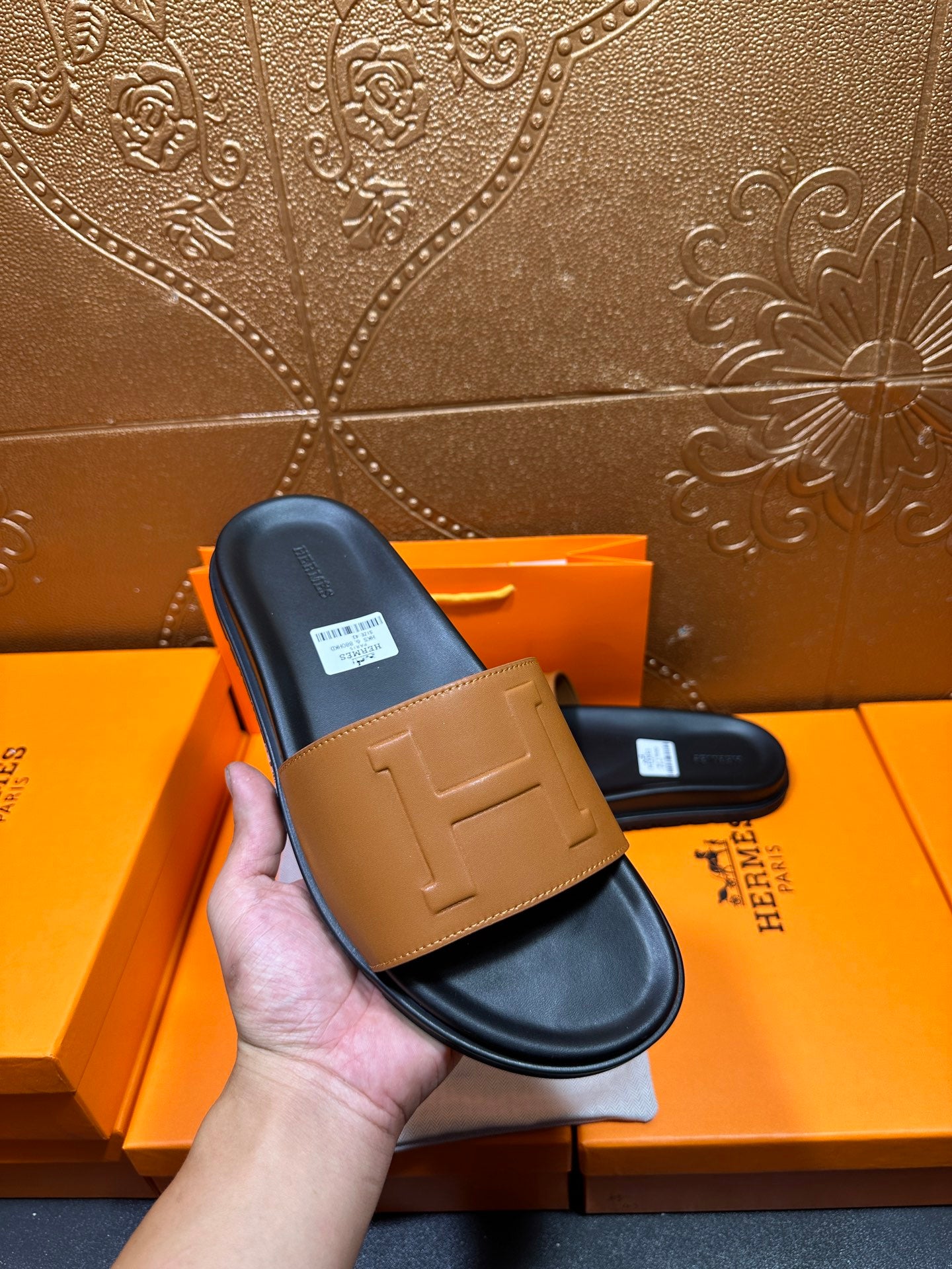 Hermes slide sandals held in hand: tan front strap on dark footbed