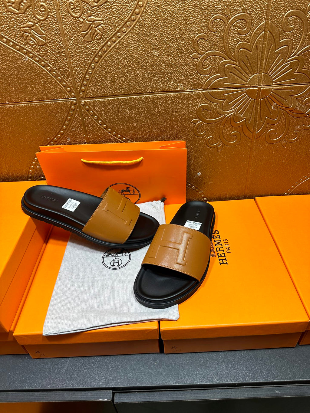 Hermes slide sandals pair with tan straps and black footbeds, on orange packaging