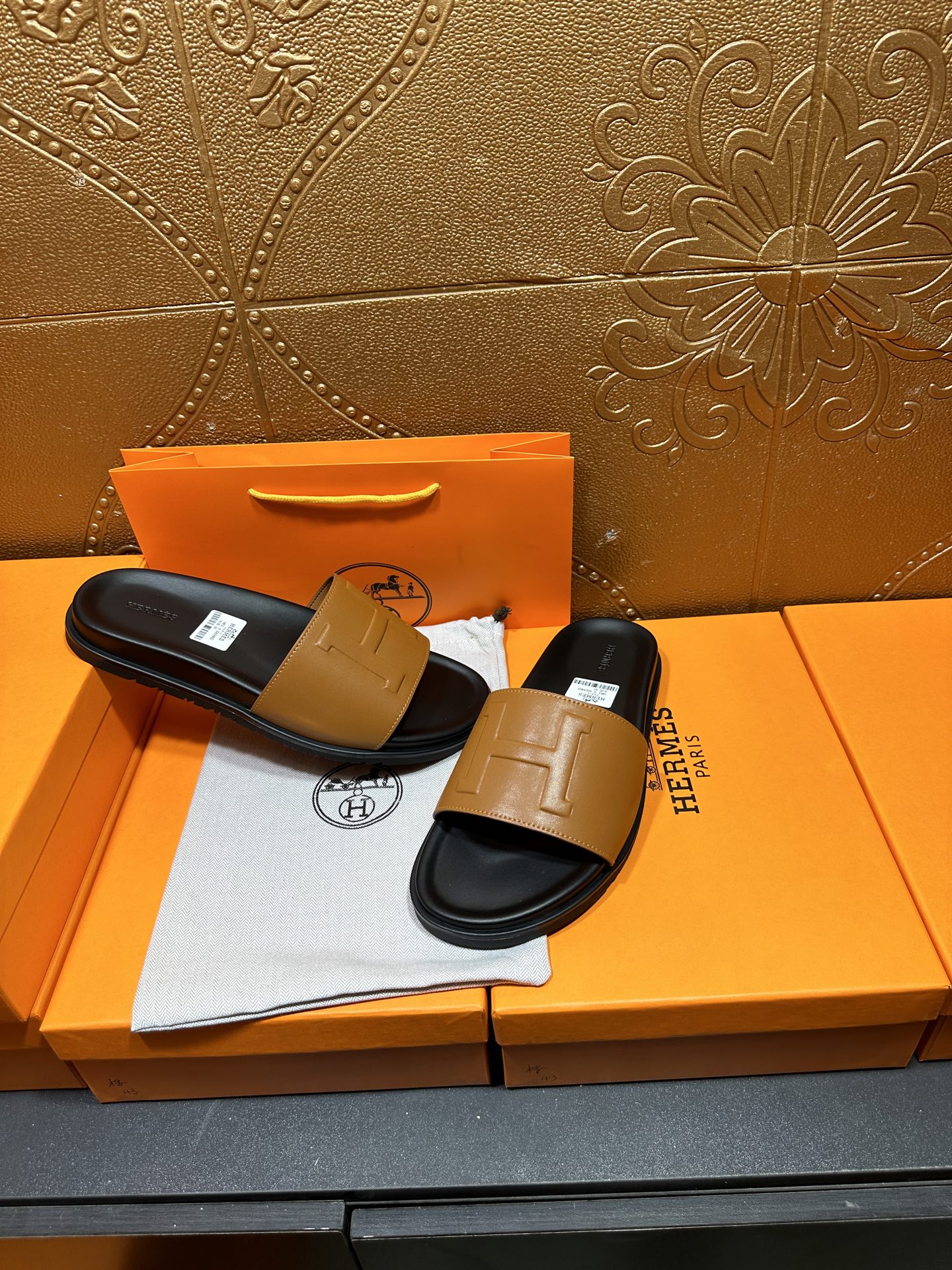 Hermes slide sandals pair with tan straps and black footbeds, on orange packaging