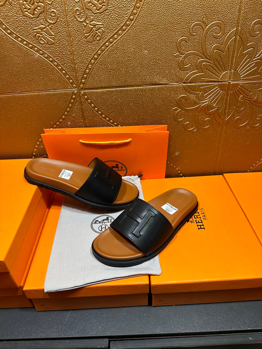 Black leather slide sandals with embossed H strap and tan footbed