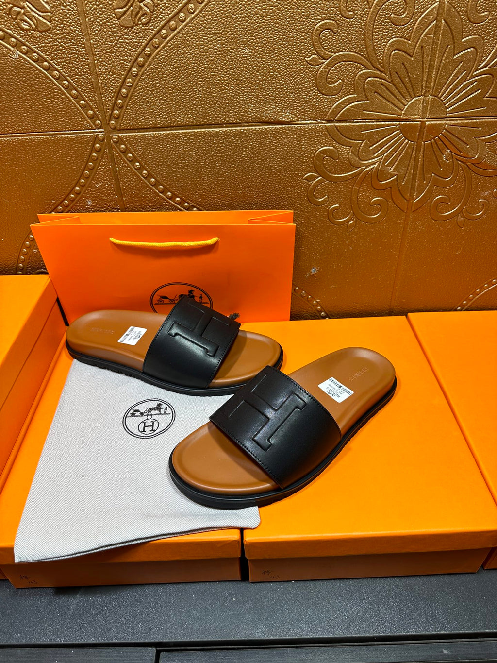 Black leather slide sandals with embossed H strap and tan footbed