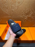 Hand holding a leather slide sandal with wide strap and H stitch detail