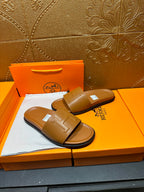 Tan leather Hermes slide sandals with square buckle strap, pair angled on box