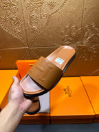 Hand holding tan leather Hermes slide sandal, square buckle strap on top