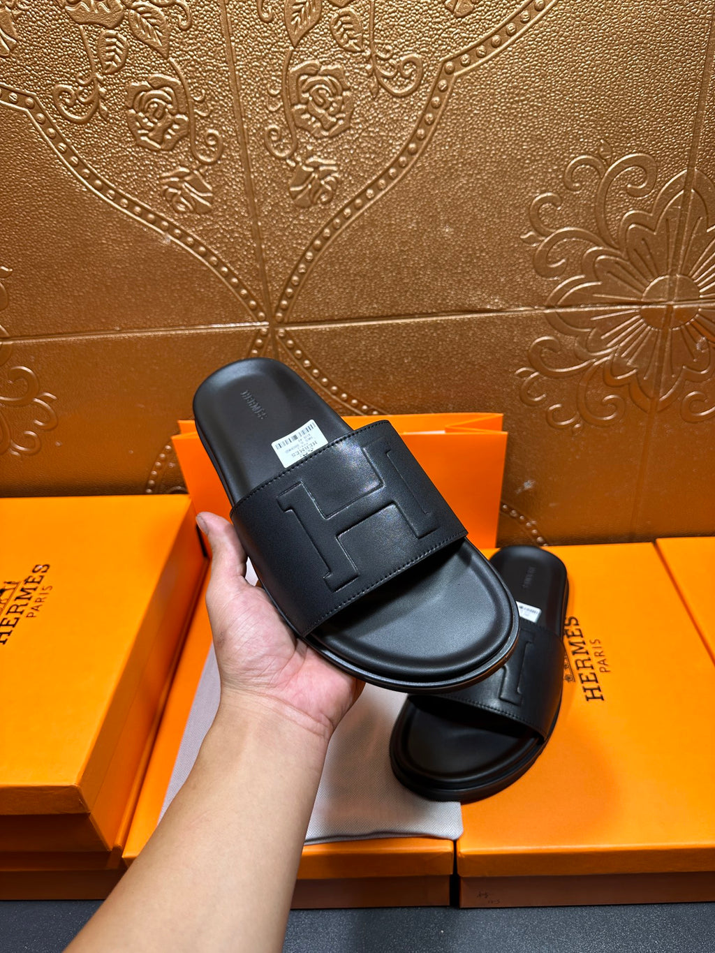 Black Hermes slide sandal held in hand, H-style strap detail visible.