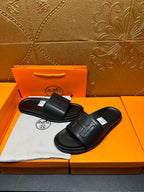 Black Hermes slide sandals with H-style strap, pair positioned on shoe box.