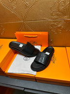 Black Hermes slide sandals with H-style strap detail, pair shown on box.