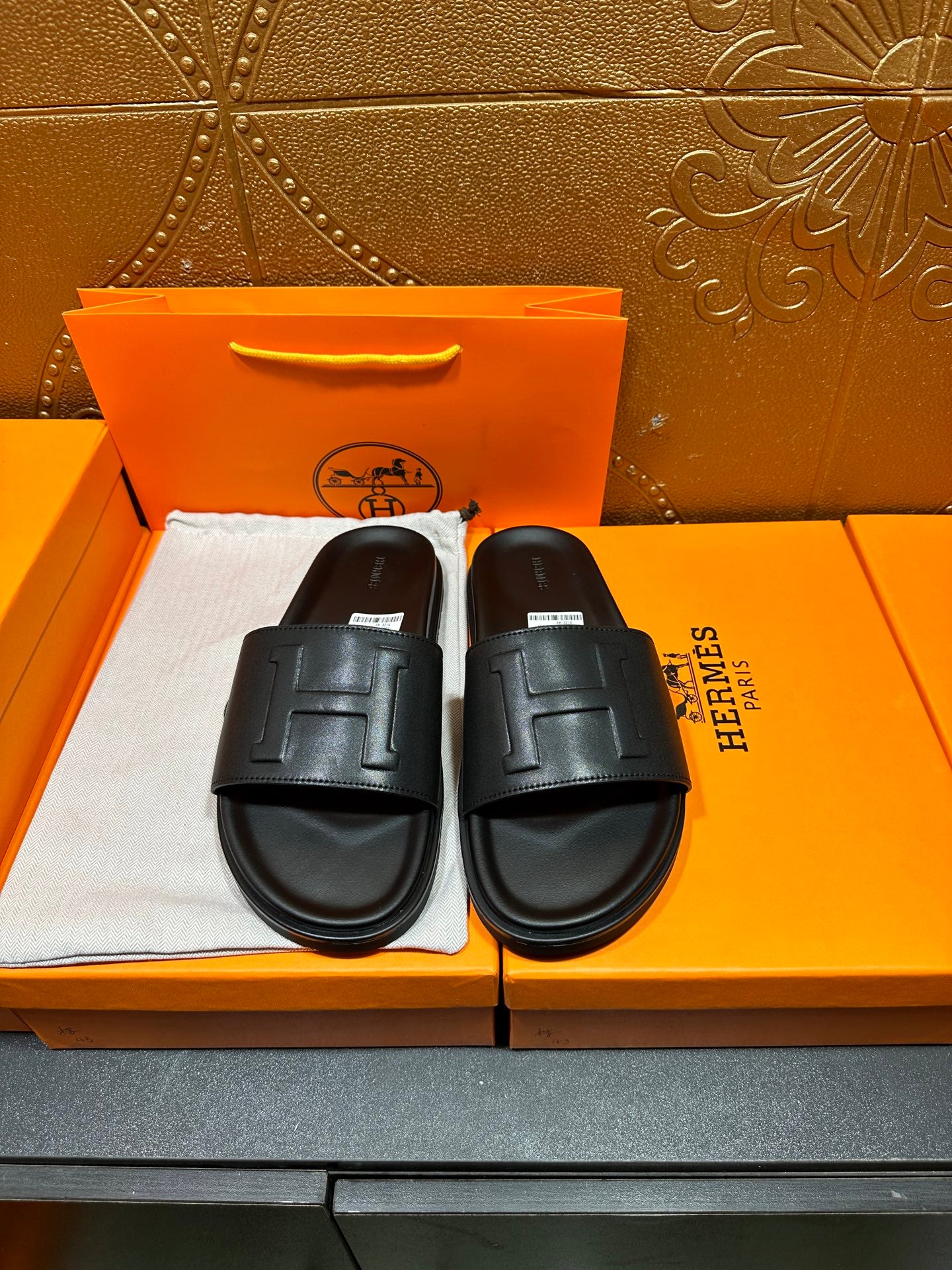 Black Hermes slide sandals with H-style strap detail, pair shown on box.