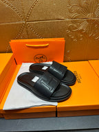 Black Hermes slide sandals with H-style strap detail, pair laid on box top.