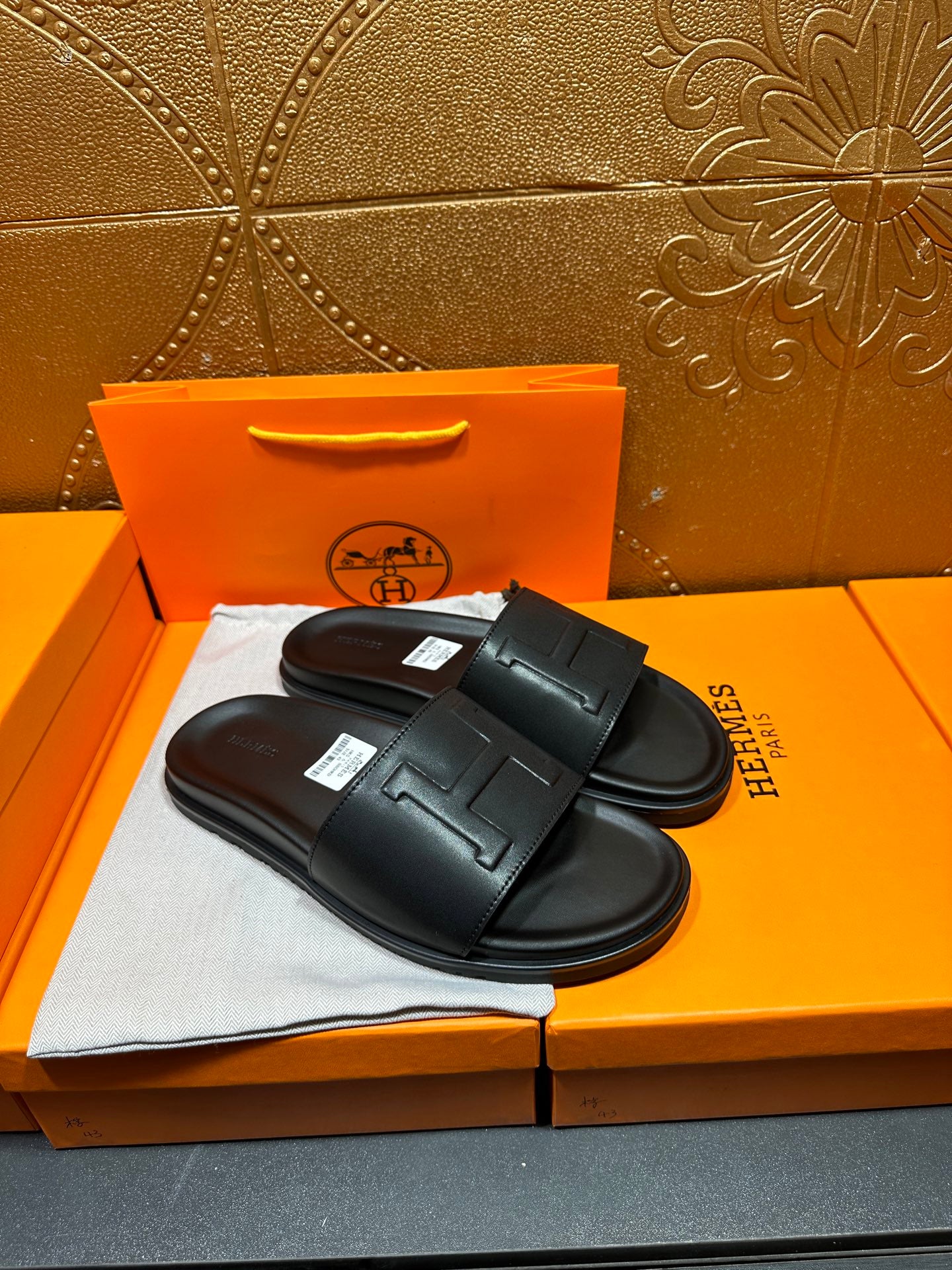 Black Hermes slide sandals with H-style strap detail, pair laid on box top.