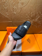 Black Hermes slide sandal held up, wide strap with H-style logo detail.
