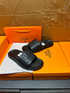 Black Hermes slide sandals with wide straps and H-style logo, pair on box.