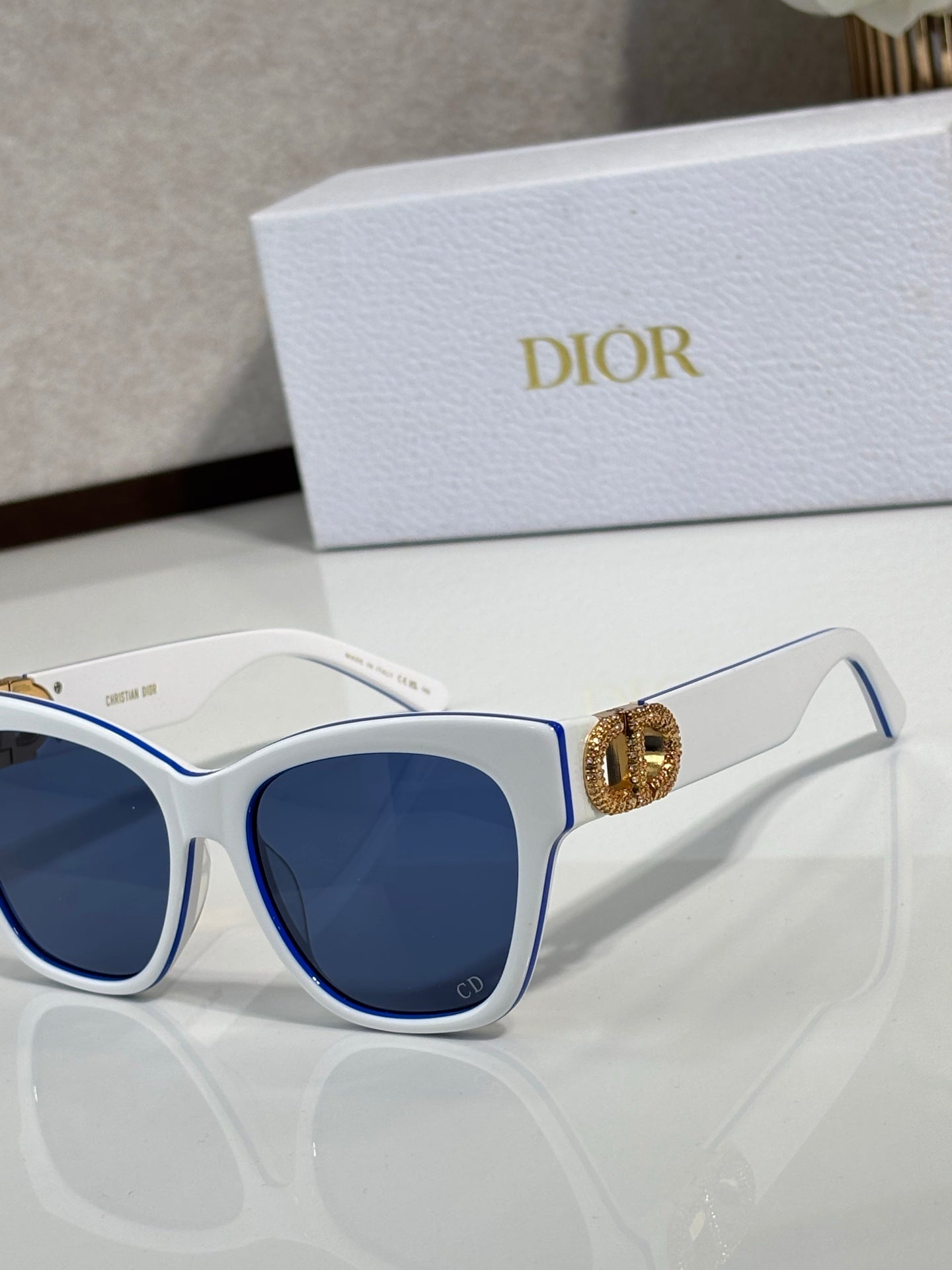 White cat-eye sunglasses close-up with blue lenses and gold logo charm detail