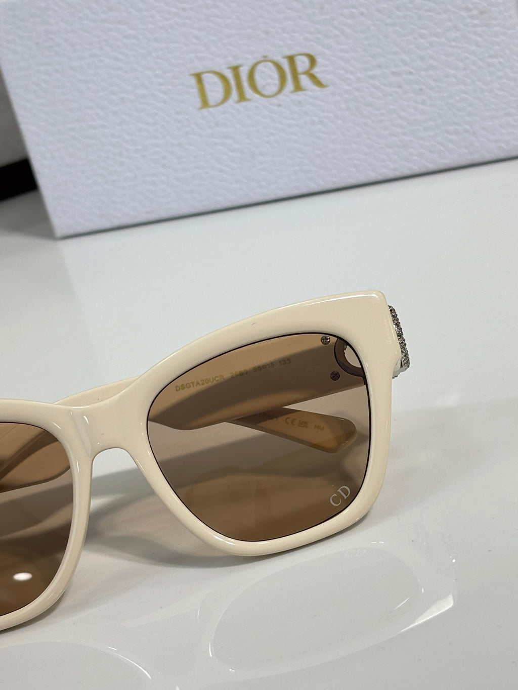 Cream cat-eye sunglasses close-up with gold logo charm and tinted lens