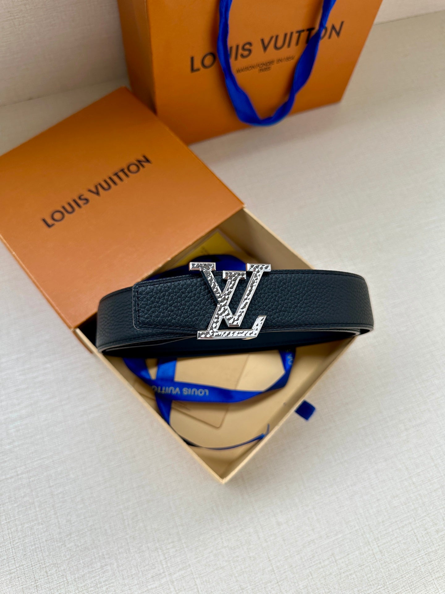 Dark blue leather belt with gold LV buckle, resting on product box and paper.
