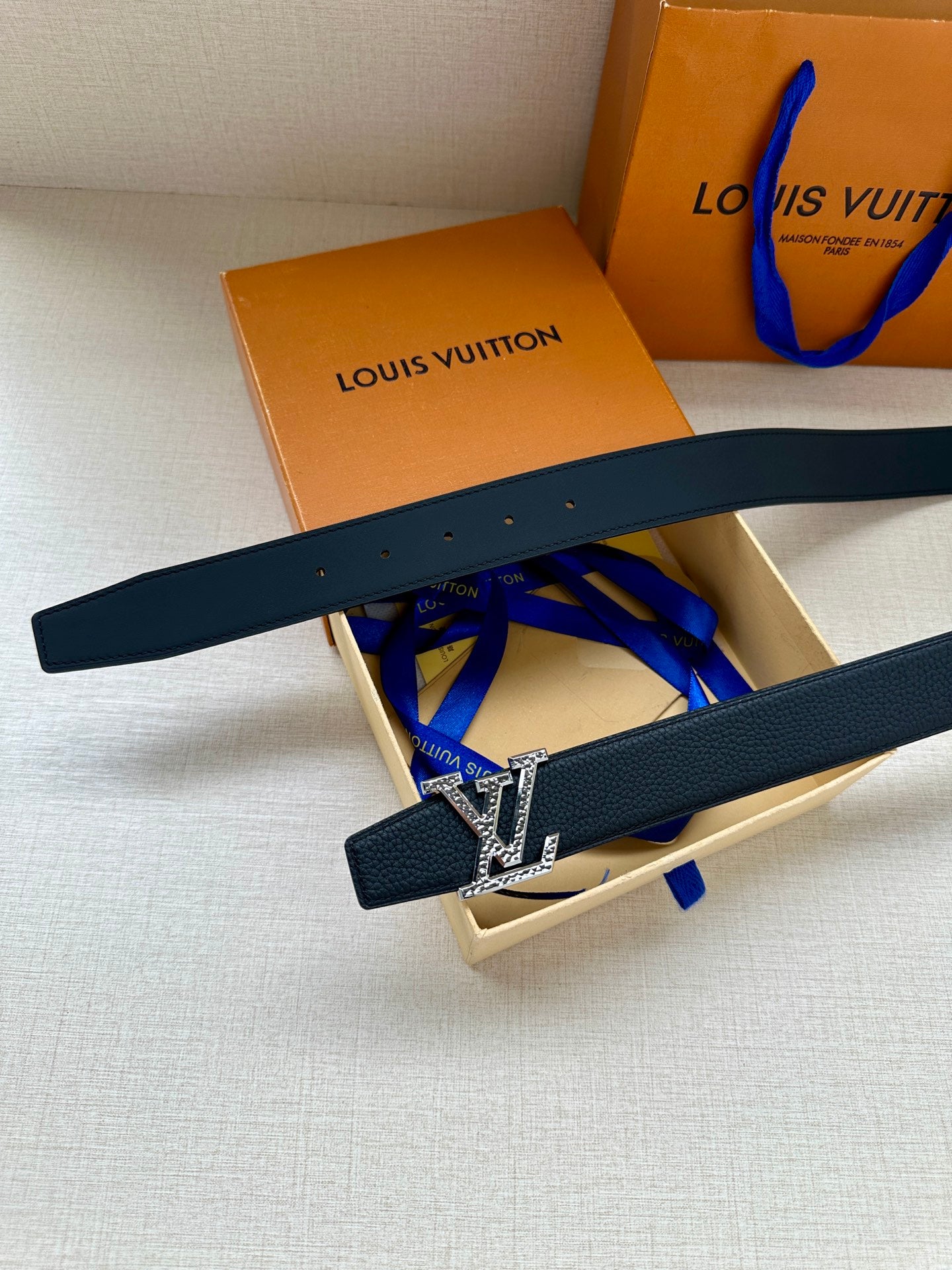 Dark blue leather belt with gold LV buckle, shown laid diagonally over box.