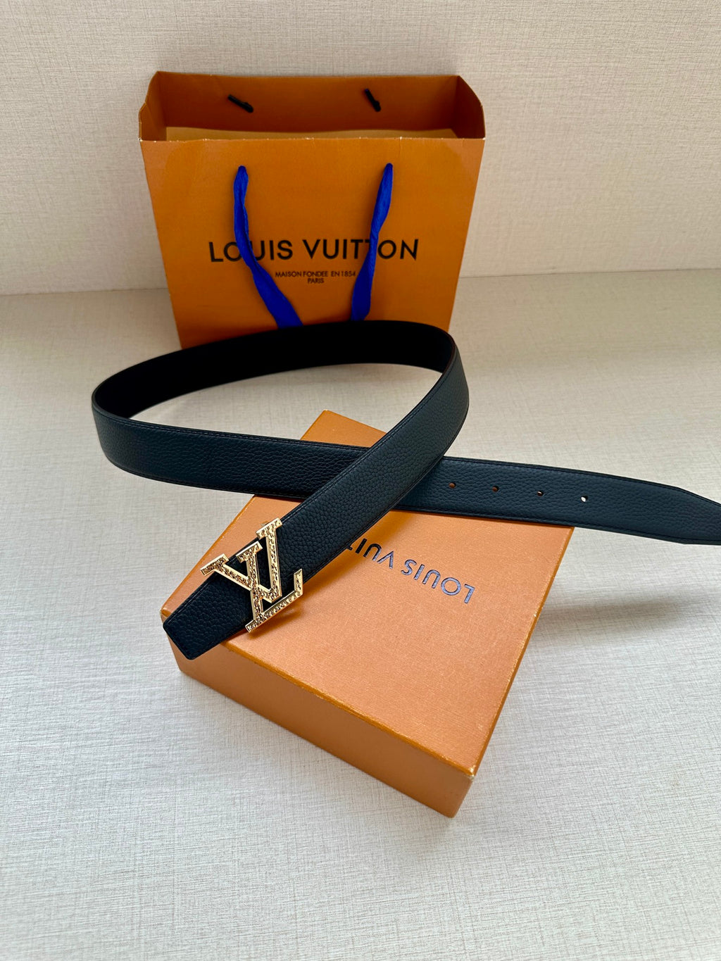 Dark blue leather belt with gold LV buckle and strap holes, laid across box.
