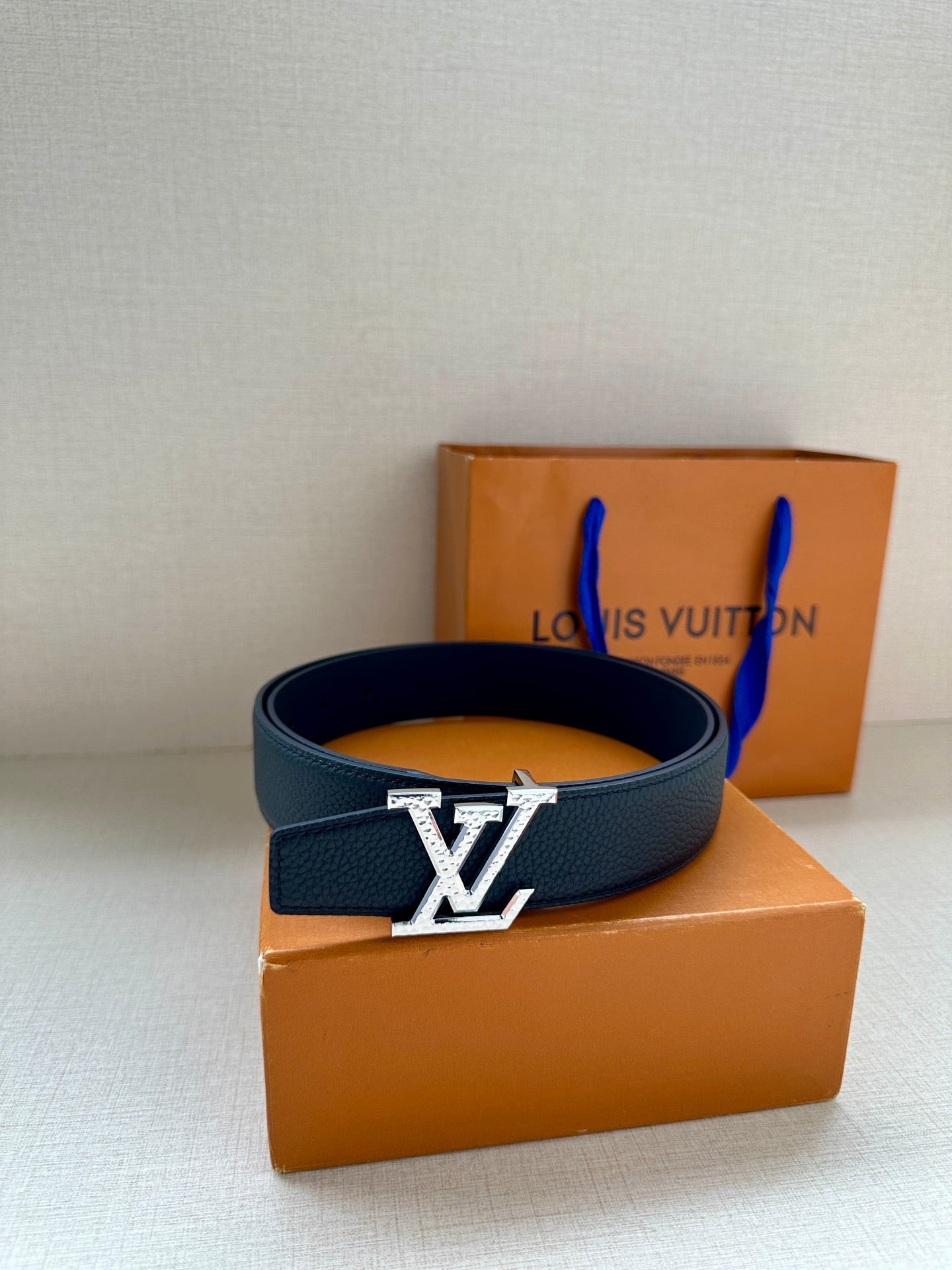 Dark blue leather belt with gold LV buckle, placed atop orange box.