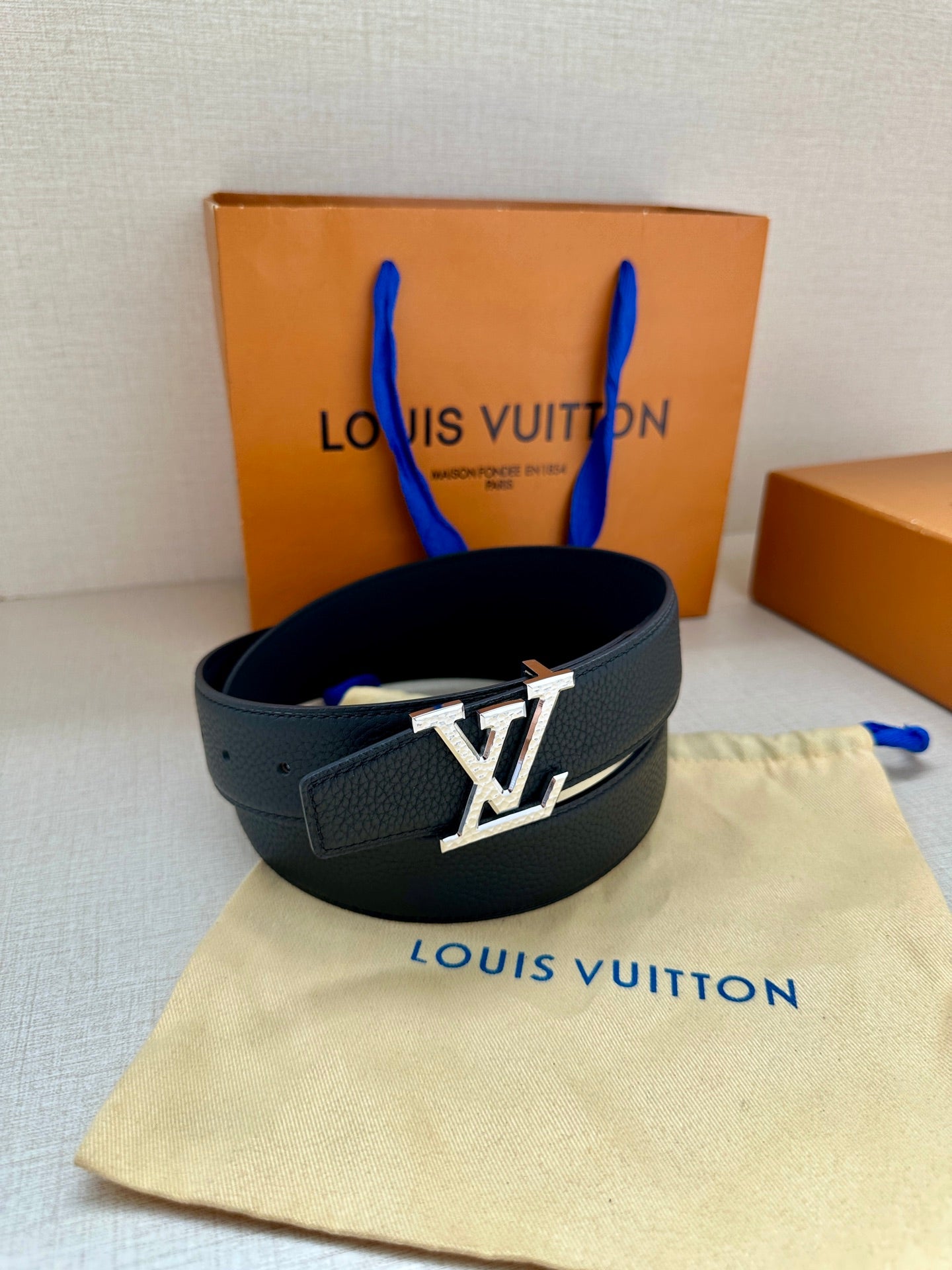 Dark blue leather belt with gold LV buckle, looped on a cloth with box behind.
