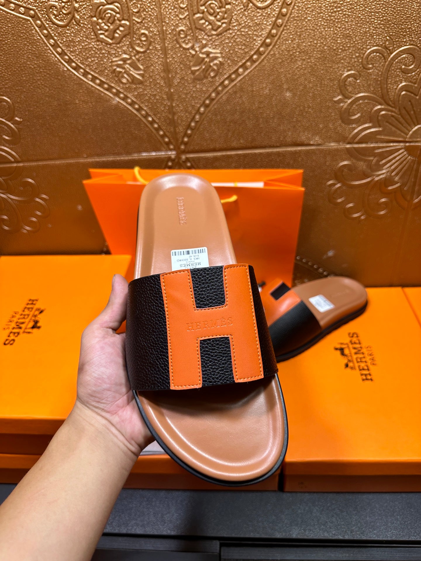 Hermes slide sandal shown in hand with black straps and orange H patch, tan footbed