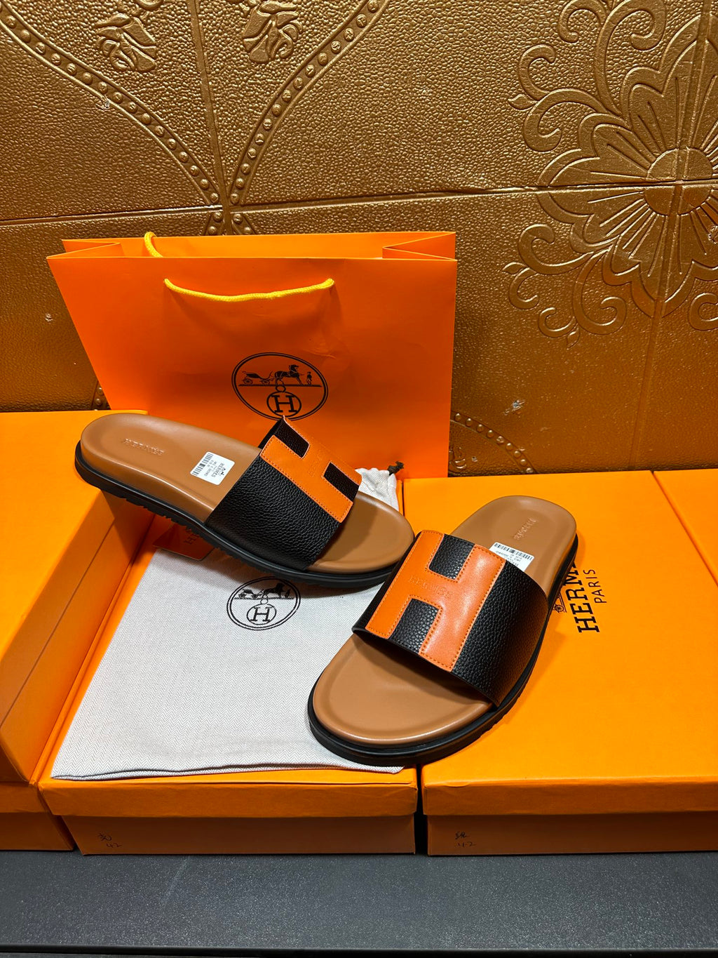 Hermes slide sandals on display: black straps with orange H detail, tan soles