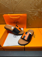 Hermes slide sandals on display: black straps with orange H detail, tan soles
