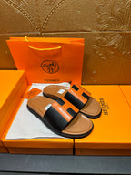 Hermes slide sandals with black-and-orange H detail across the strap, tan sole