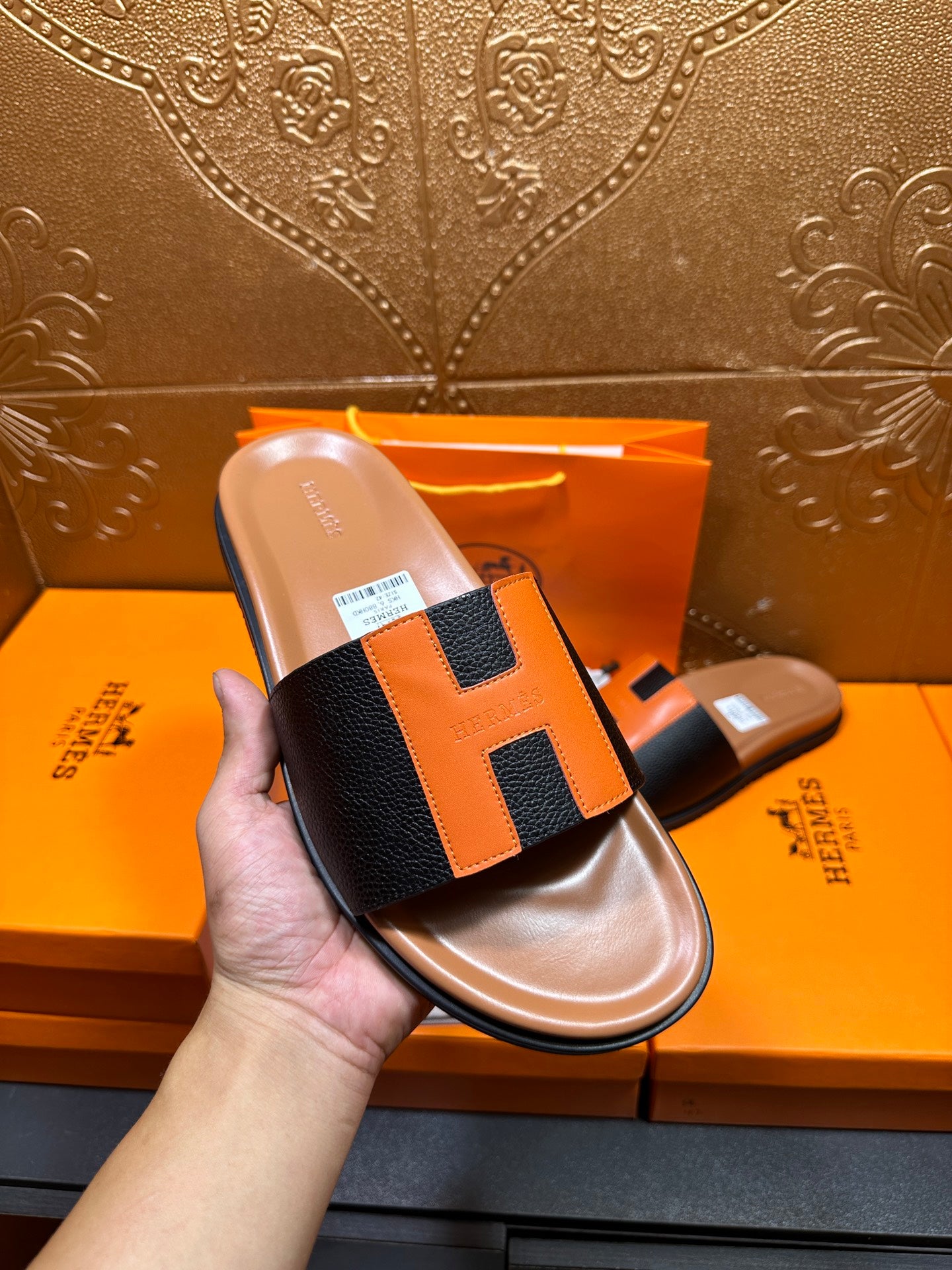 Hermes slide sandal shown in hand: black upper with orange H detail, tan sole