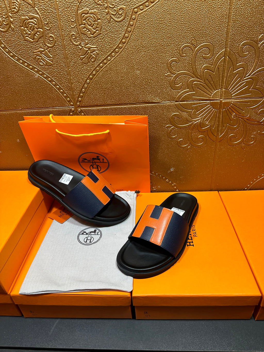Two black slide sandals with orange H strap detail on a cloth insert