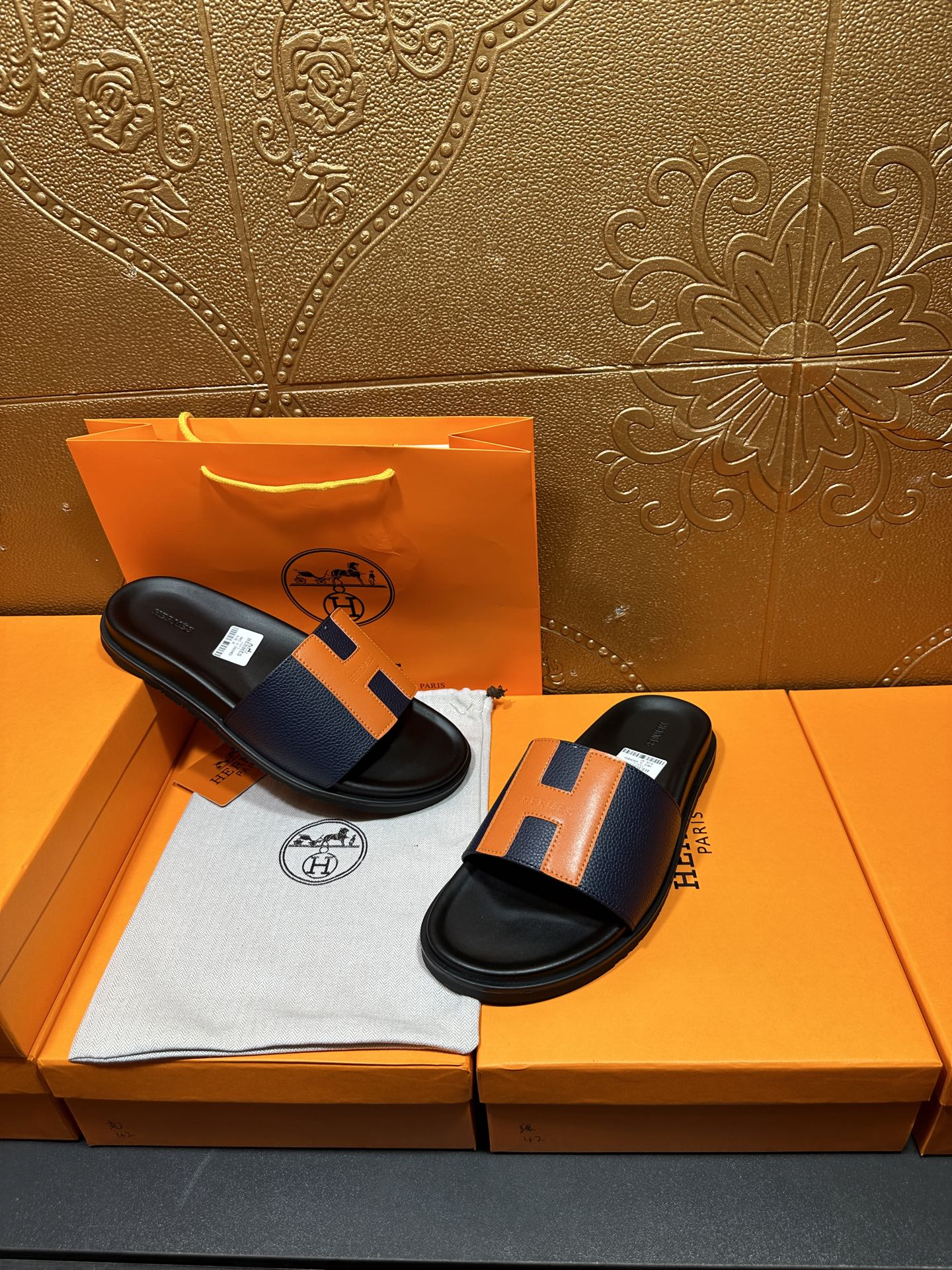 Two black slide sandals with orange H strap detail on a cloth insert