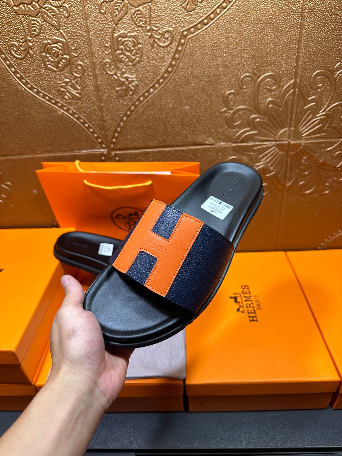 Hand holding black slide sandal with orange H strap detail