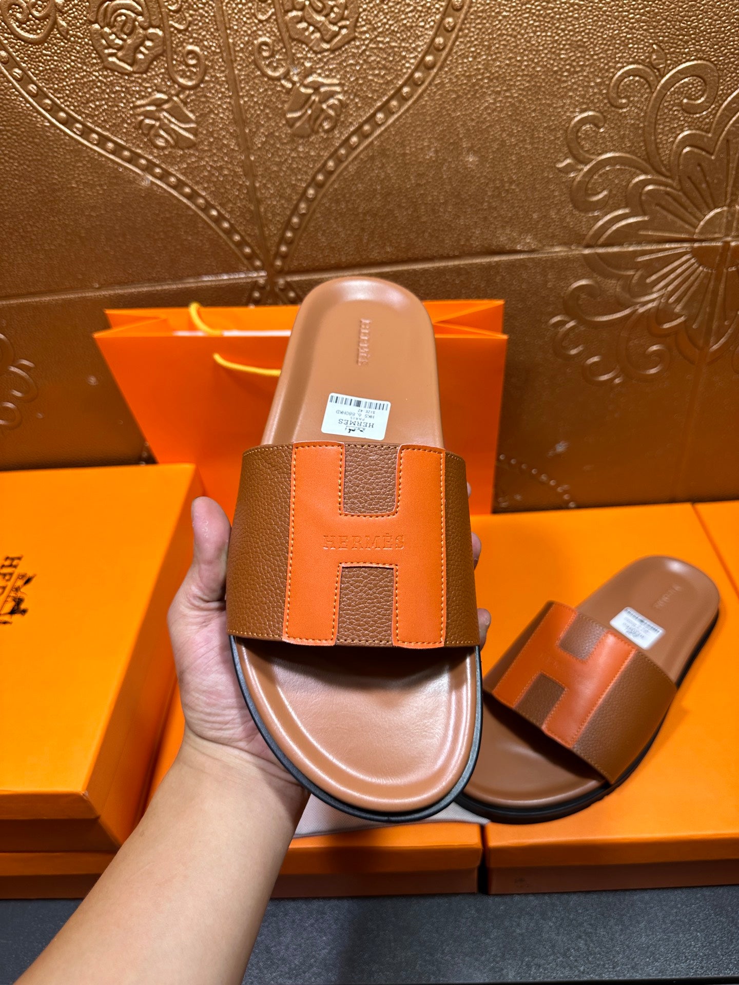 Hand holding tan slide sandal with orange square H strap and black outsole.