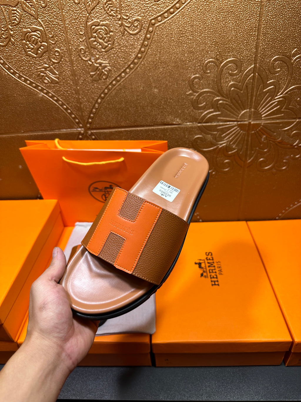 Hand holding tan H-strap slide sandal with orange panels above open box.