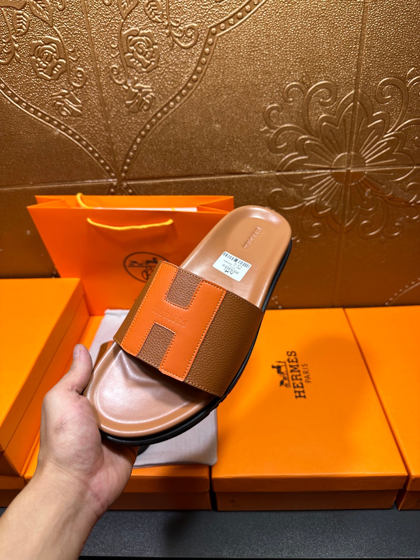 Hand holding tan H-strap slide sandal with orange panels above open box.