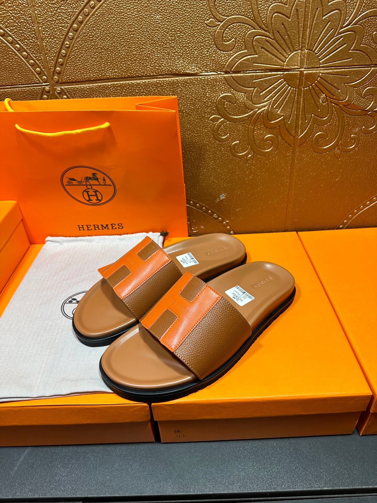 Tan H-strap slide sandals with orange panels and black outsole, on box padding.