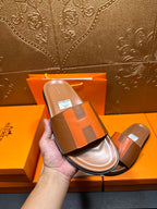 Hand holding tan slide sandals with orange H strap and black-trim sole.